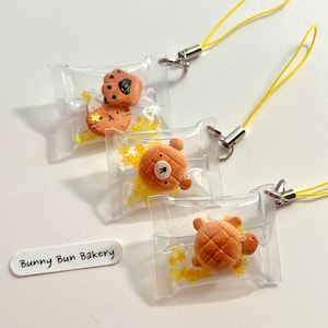 Könnte beinhalten: Drei kleine, transparente Plastiktüten mit gelbem Sternenglitzer im Inneren. Jede Tüte enthält ein anderes tierförmiges Gebäck: eine Katze, einen Bären und eine Schildkröte. Die Tüten sind an gelben Schnur-Lanyards befestigt. Der Text "Bunny Bun Bakery" steht auf einem weißen Etikett.