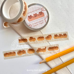 May include: A roll of washi tape with a design of cartoon cats and bread loaves. The tape is labeled "Bunny Bun Bakery Fresh Loafs Washi Tape".