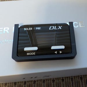 May include: A black electronic device with the text "SOLER PRF. DLX" and "MODE" in white. The device has two rectangular buttons and two arrow symbols. It is set on a white surface with the word "PERFORMANCE" visible.