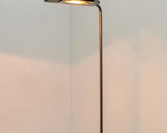 May include: A tall, slender floor lamp with a dark metal finish. The lamp features a circular base, a long, straight pole, and a flat, oval-shaped light fixture at the top. The light is on, illuminating the area.
