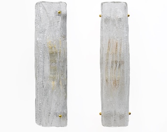 May include: Two clear, textured glass wall sconces. Each sconce is rectangular with a rough, bark-like texture and small gold-coloured accents. The sconces are designed to hold a light bulb, creating a warm glow.