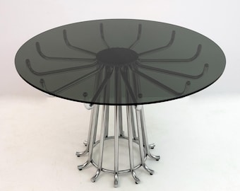 May include: A round, modern dining table with a smoked glass top and a chrome base. The base features a series of vertical chrome bars that curve outward at the bottom. The table's design is sleek and minimalist, suitable for a contemporary interior.