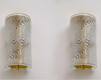 May include: Two cylindrical wall sconces with a textured, swirling pattern in silver and gold. The sconces have a clear, rounded base and a gold-coloured light fixture. The sconces are mounted on a white wall.