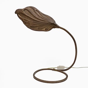 Può includere: Lampada da tavolo color bronzo con paralume a forma di foglia. Il paralume ha una superficie strutturata con linee curve. Lo stelo della lampada si curva graziosamente verso una base circolare. La lampada è collegata a una presa bianca.
