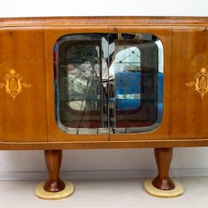 May include: A vintage wooden cabinet with a glossy finish. The cabinet features a central glass-door section and decorative gold accents. It stands on four curved legs with circular bases, showcasing a classic design.