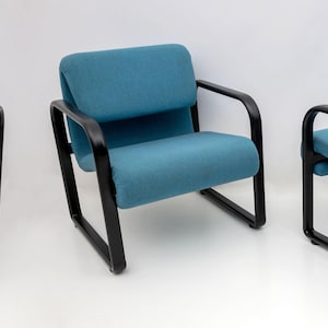 May include: Three teal armchairs with black metal frames are shown against a white backdrop. The chairs have a boxy design with a padded seat and back. The armrests are curved and the legs are straight.