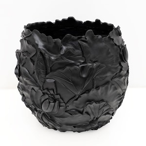 May include: A black, round decorative planter with a detailed relief of lotus flowers and leaves. The planter has a textured surface and a wide opening. The design is carved into the exterior of the planter.