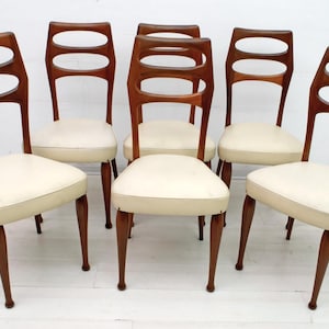 May include: Six mid-century modern dining chairs. The chairs have a dark wood frame with a curved back featuring three oval cutouts. The seats are upholstered in a cream-colored fabric. The chairs have tapered legs and a classic design.