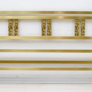 Può includere: Una struttura letto in metallo color oro con una testiera rettangolare. La testiera presenta tre pannelli verticali decorativi con dettagli strutturati. La struttura ha un design a barre orizzontali ed è realizzata in un materiale metallico lucido.