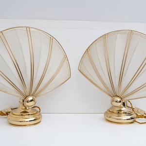 Può includere: Due lampade da tavolo a forma di ventaglio abbinate. Ogni lampada presenta una base e una cornice color oro, che sostengono un paralume bianco sporco semitrasparente. Le lampade hanno un design vintage, adatto all'arredamento.