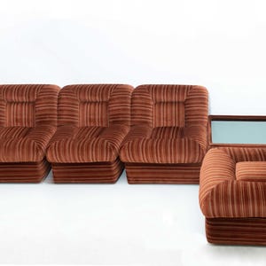 May include: A set of modular seating in a warm brown color. The set includes five individual seat sections and one armchair, all with a corduroy-like texture. The design is a retro style, with a low profile and boxy shape.