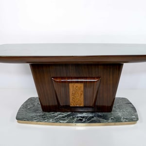 May include: A mid-century modern style table with a dark wood base and a light-colored top. The base has a geometric design with a decorative wood panel. The table sits on a marble base with brass trim.
