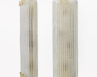 May include: Two clear glass wall sconces with a textured, ribbed design. Each sconce has a rectangular shape with gold-coloured metal accents at the top and bottom. The sconces are designed to be mounted on a wall and provide ambient lighting.