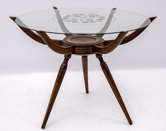 May include: A vintage, three-legged side table with a round glass top. The table has a dark brown wooden frame with curved supports and decorative floral etching on the glass. The table is approximately 24 inches in height.