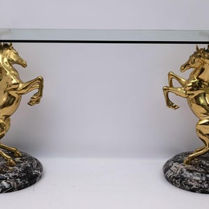 Könnte beinhalten: Ein goldfarbener Konsolentisch mit rechteckiger Glasplatte. Der Tisch wird von zwei aufsteigenden Pferdeskulpturen getragen. Die Pferde stehen auf ovalen Sockeln aus schwarz-weißem Marmor.