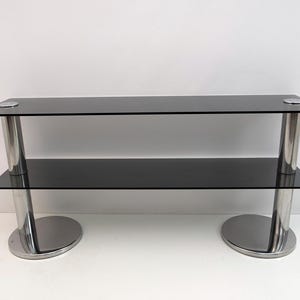 May include: A modern two-tier console table with a sleek design. The table features two black glass shelves supported by polished chrome legs. The legs are cylindrical and connect to circular bases. The top shelf has two small, round, silver accents.