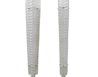May include: Two clear glass wall sconces with a textured, leaf-like pattern. Each sconce has a rectangular shape with a tapered design and small gold-coloured accents. The sconces are designed for wall mounting and are ideal for ambient lighting.