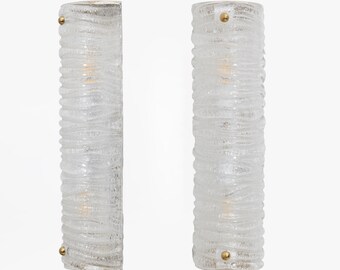 May include: Two clear glass wall sconces with a textured, wavy design. Each sconce has a cylindrical shape with a brass-coloured accent at the top and bottom. The sconces are illuminated, suggesting they are functional lighting fixtures.