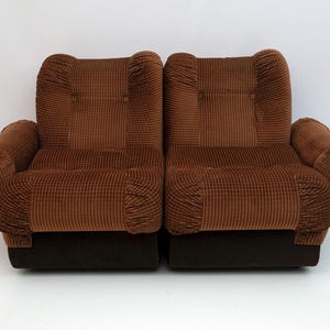 May include: Two brown corduroy armchairs with rounded arms and backs. The chairs have a dark brown base and a textured, striped pattern. The chairs are side by side, creating a symmetrical design.