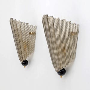 May include: Two matching wall sconces with a fan-like design. The sconces are made of ribbed, translucent glass in a smoky gray color. Each sconce has a black, spherical base and a small gold-colored accent. The sconces are mounted on a white wall.