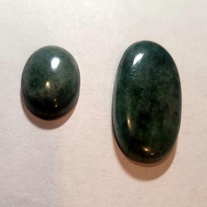 May include: Two polished, oval-shaped gemstones. One is smaller and more rounded, the other is larger and elongated. Both stones have a deep green color with subtle variations in tone, and a glossy finish.