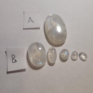 May include: A collection of seven oval-shaped moonstone cabochons in various sizes. The stones are white with a slight iridescent sheen. The stones are arranged in a row, with the largest stone at the top and the smallest stones at the bottom.