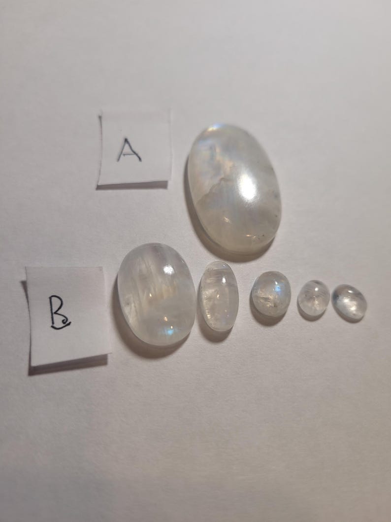 May include: A collection of seven oval-shaped moonstone cabochons in various sizes. The stones are white with a slight iridescent sheen. The stones are arranged in a row, with the largest stone at the top and the smallest stones at the bottom.