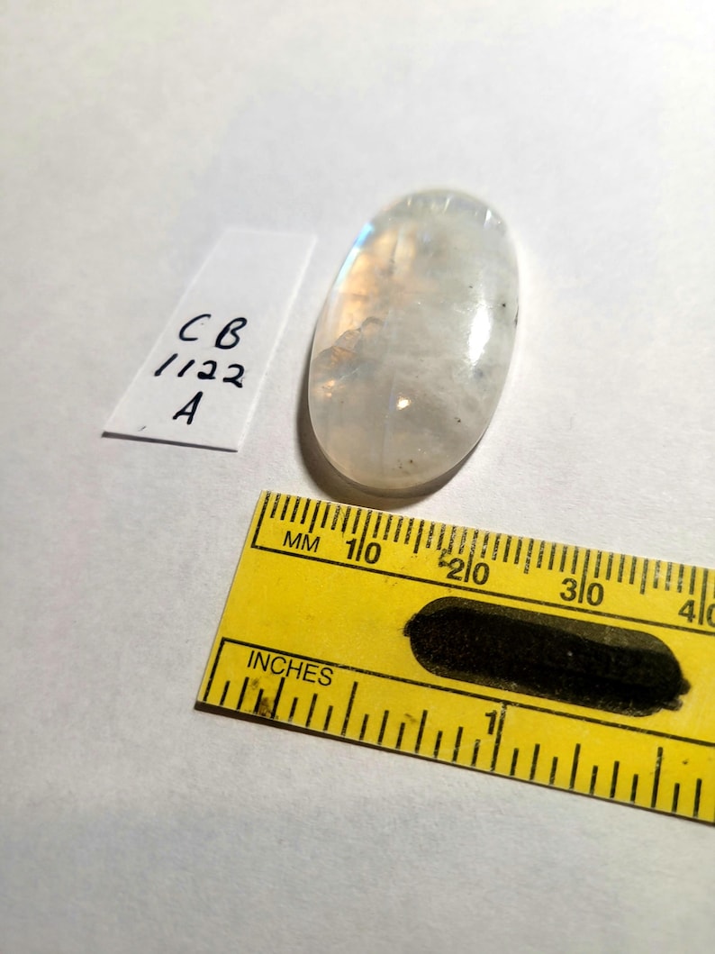 May include: A white oval moonstone cabochon with a slight rainbow sheen. The stone is approximately 1 inch long and 1/2 inch wide. The stone is sitting on a white surface next to a ruler.