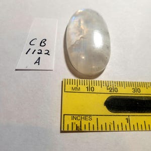 May include: A white oval moonstone cabochon with iridescent rainbow colors. The stone is approximately 1 inch long and 1/2 inch wide. The stone is sitting on a white surface next to a ruler.