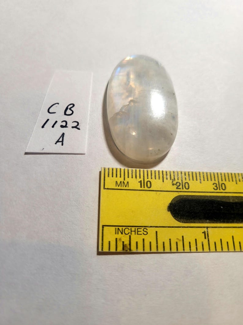 May include: A white oval moonstone cabochon with iridescent rainbow colors. The stone is approximately 1 inch long and 1/2 inch wide. The stone is sitting on a white surface next to a ruler.