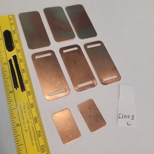 May include: Eight rectangular metal tags with a hole in the center. The tags are various shades of copper and have a slightly textured surface. The tags are arranged in a random pattern on a white surface. A ruler is visible in the bottom left corner of the image. A piece of paper with the text "C1003" is visible in the bottom right corner of the image.