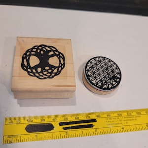 Puede incluir: Dos sellos de madera con diseños negros. El primer sello es cuadrado y presenta un diseño de árbol de la vida. El segundo sello es redondo y presenta un diseño de flor de la vida.