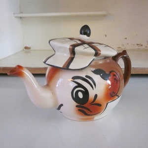 May include: A white ceramic teapot with a brown and white striped lid. The teapot is painted with a cartoon face with brown eyes, a brown nose, and a brown mustache.