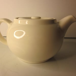 Puede incluir: Tetera de cerámica color crema con cuerpo redondeado, asa curva y boquilla. La tapa tiene un pequeño pomo para facilitar su extracción. La tetera está diseñada para preparar y servir té. El acabado brillante refleja la luz, resaltando su superficie lisa.