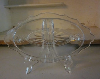May include: A clear glass serving dish with a scalloped edge and a raised, floral design in the center. The dish is on a clear plastic stand with three legs.