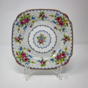May include: Square white plate with a floral cross-stitch design. The plate features pink, blue, and yellow flowers with a gold trim. The plate is displayed on a clear plastic stand.