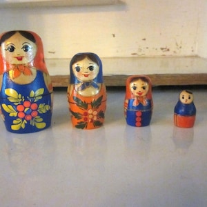 Peut inclure: Ensemble de quatre poupées russes, ou poupées gigognes, alignées. Les poupées sont peintes avec des couleurs vives, notamment le bleu, l'orange et le rouge. La plus grande poupée mesure environ 13 cm de haut.