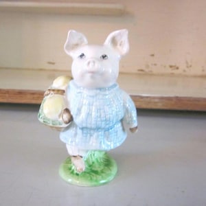 May include: A small porcelain figurine of a pig wearing a blue sweater and carrying a basket of yellow eggs. The pig is standing on a green base.