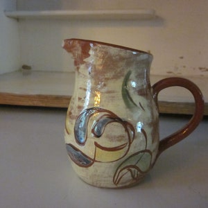 May include: A small, hand-painted ceramic jug with a brown handle and a cream base. The jug is decorated with a colourful floral design in shades of blue, green, yellow, and brown.