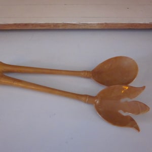 Puede incluir: Un juego de dos utensilios de servir ensalada de madera. La cuchara de servir es marrón y el tenedor de servir es marrón con un diseño en forma de flor.