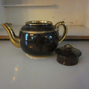 May include: A brown teapot with a gold trim and a matching lid. The teapot has a round body and a spout on the side. The lid is separate and has a knob on top.