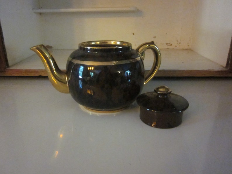 May include: A brown teapot with a gold trim and a matching lid. The teapot has a round body and a spout on the side. The lid is separate and has a knob on top.