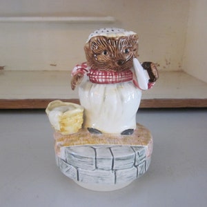 Puede incluir: Figura de cerámica de un erizo vestido con un delantal blanco y una bufanda a cuadros rojos y blancos. El erizo sostiene un pequeño paño blanco y una cesta. La figura está sobre una base de ladrillo.
