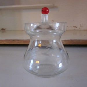 May include: A clear glass jar with a red knob on the lid. The jar has an etched floral design.