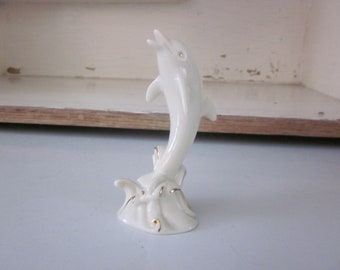 May include: A white porcelain figurine of a dolphin leaping out of the water. The dolphin is detailed with gold accents and is set on a base with a wave design.