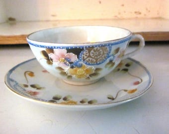 May include: A white teacup and saucer set with a floral design. The cup has a blue and gold trim and the saucer has a blue trim. The flowers are pink, yellow, and blue.