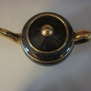 May include: A black teapot with gold trim and a gold knob on the lid. The teapot is sitting on a white surface.