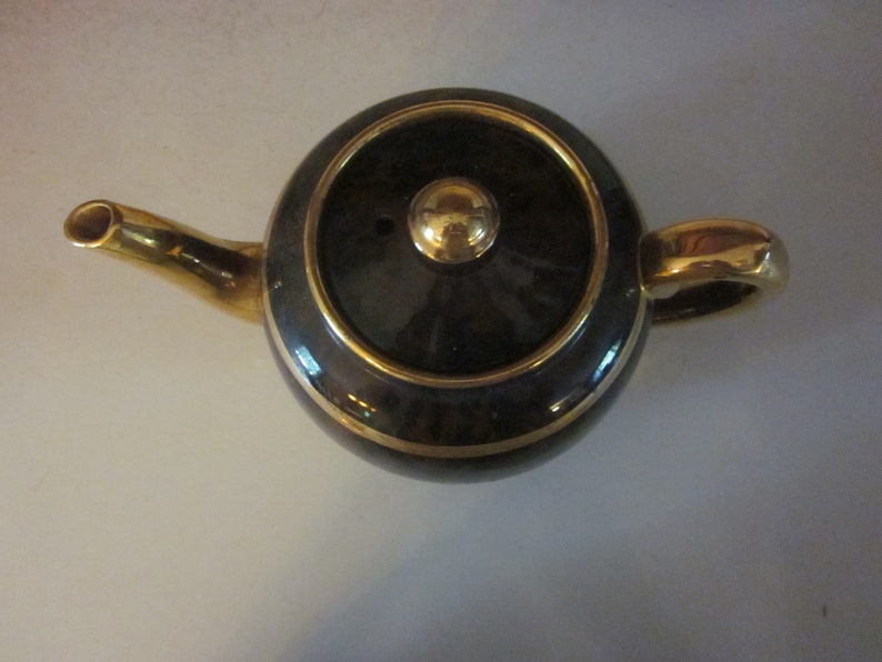 May include: A black teapot with gold trim and a gold knob on the lid. The teapot is sitting on a white surface.