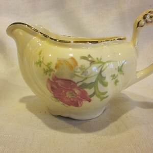 Peut inclure: Crémier en porcelaine crème avec un bord et une anse dorés. Le crémier présente un motif floral avec des fleurs roses et oranges et des feuilles vertes. La anse est ornée d'un motif floral.