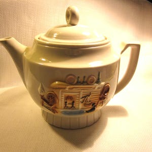 May include: A white ceramic teapot with a brown and gold design of a building with a chimney and a window. The teapot has a lid with a knob on top.